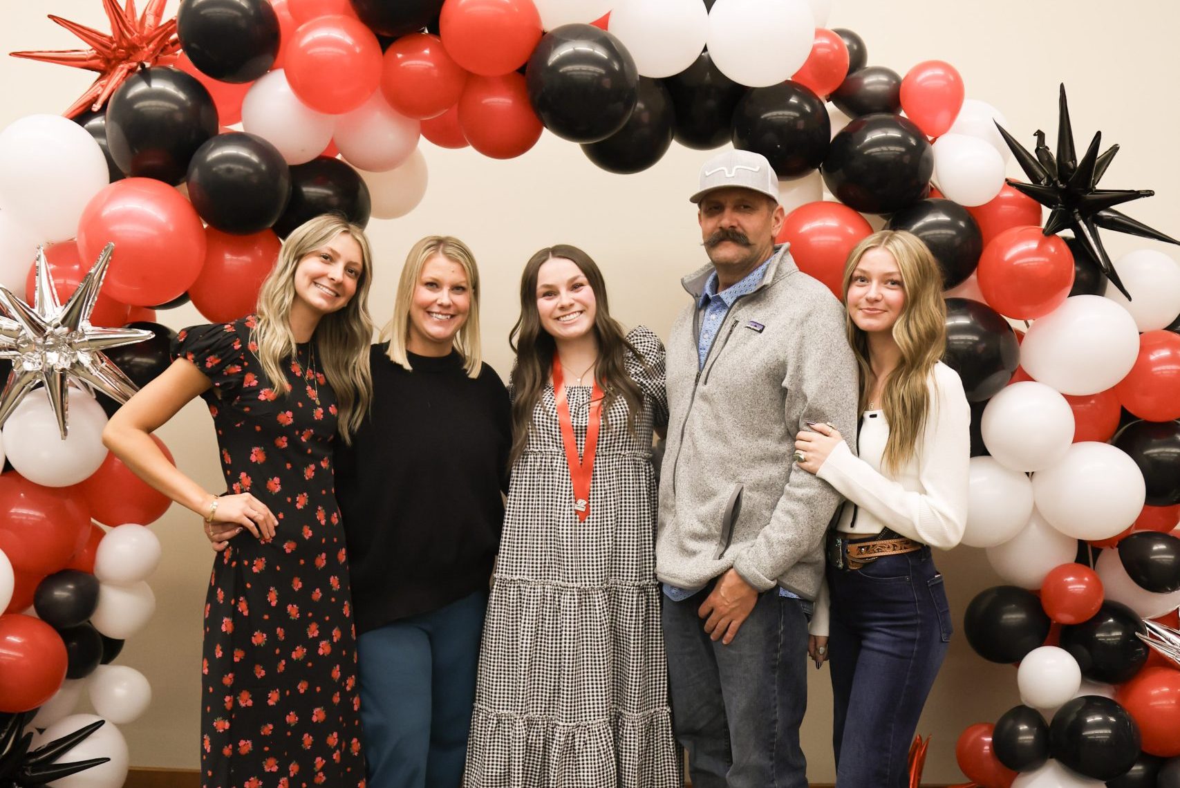 Practical nursing graduate poses for a picture with family in front of a red and black balloon arch.