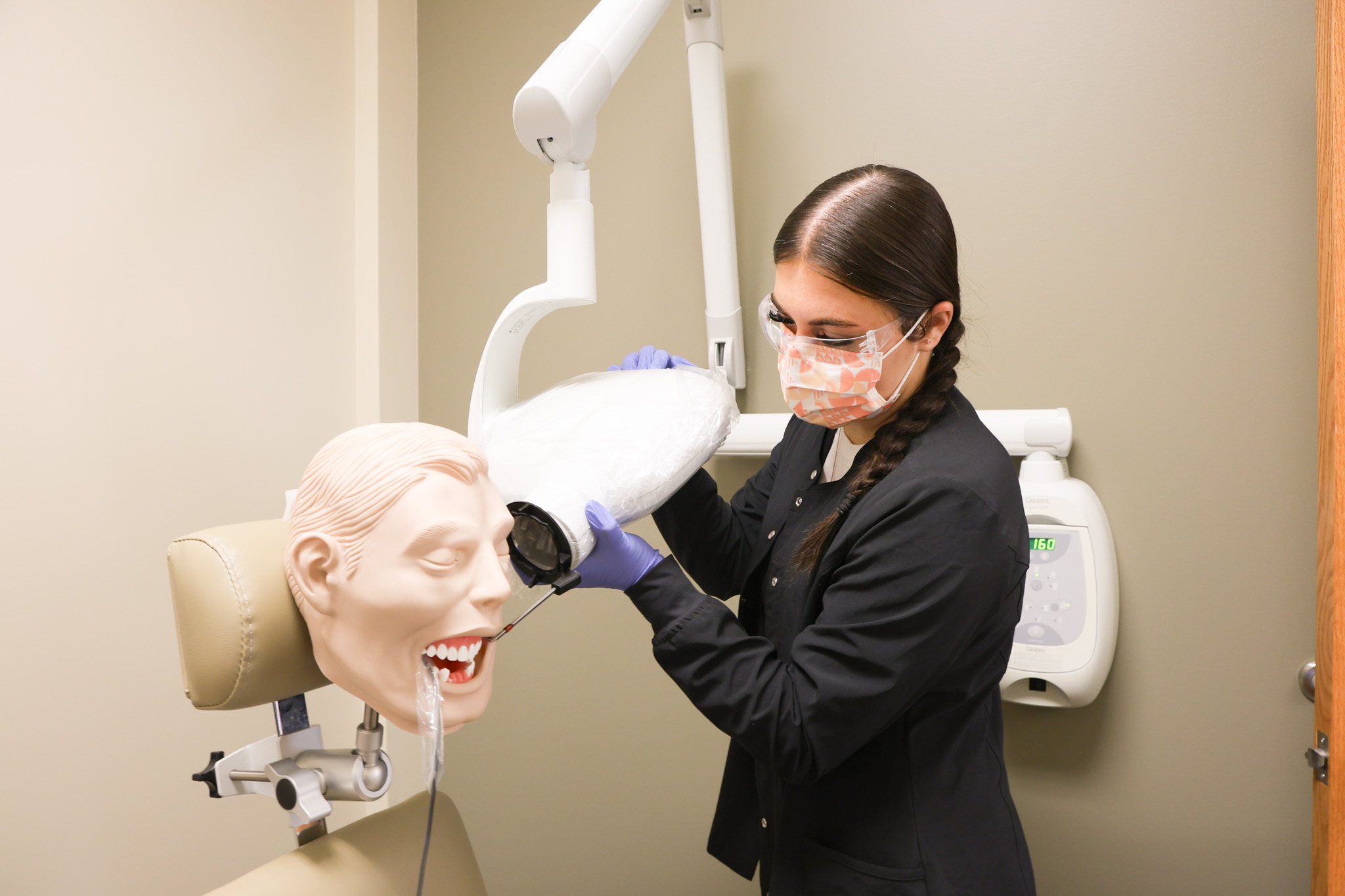 A person wearing protective gear adjusts a dental light while practicing a procedure on a dental mannequin in a clinical exam room.
