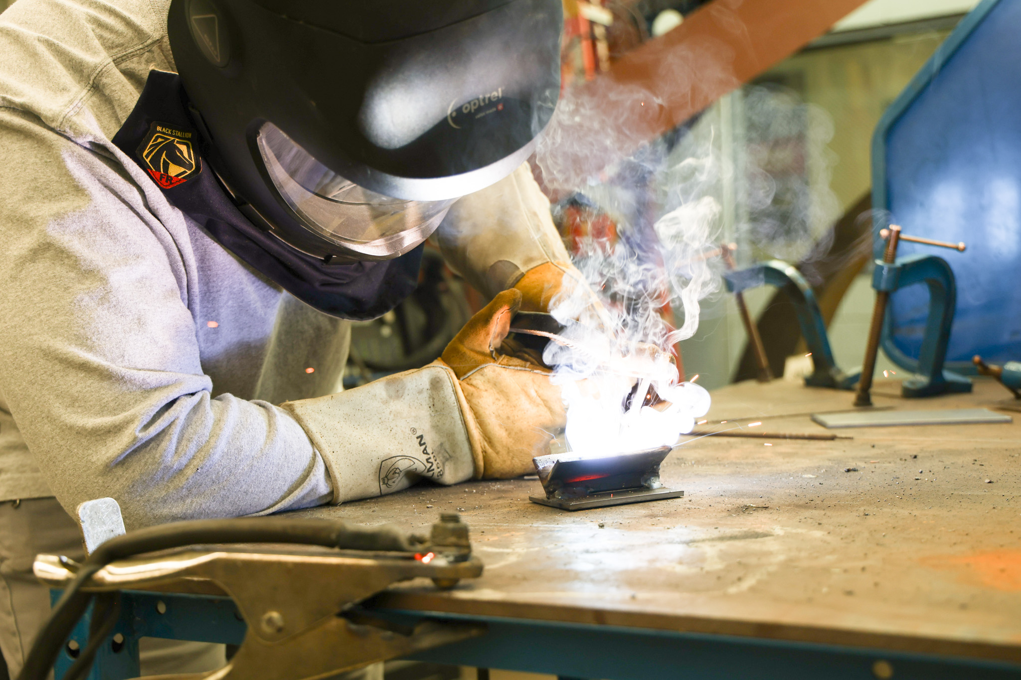 Man wearing a welding helmet and safety gear welds a piece of metal. The metal looks hot, surrounded by sparks and smoke.