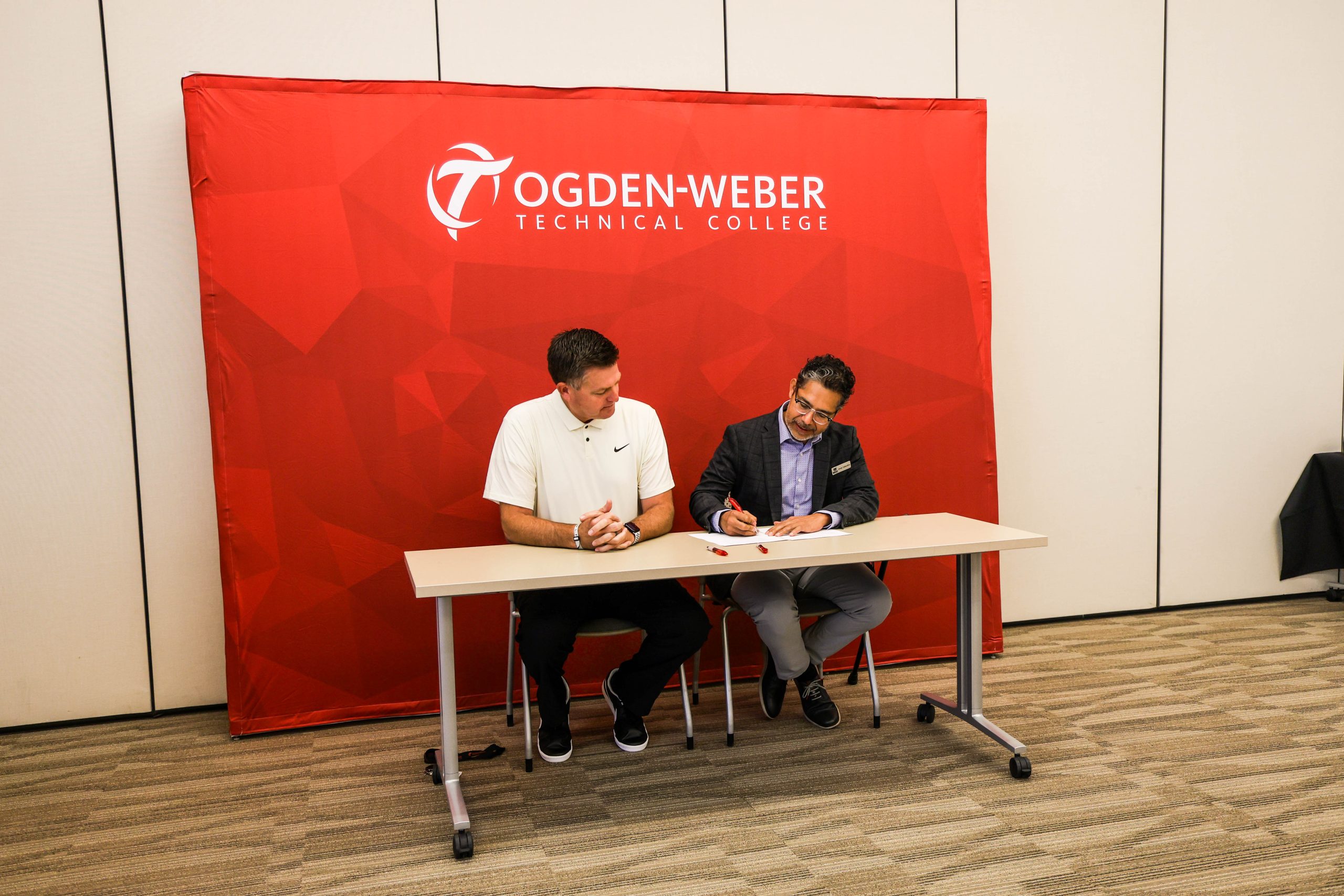 Composites_hero_video - Ogden Weber Technical College Two individuals seated at a table in front of a red Ogden-Weber Technical College backdrop. One person is writing on a document while the other looks on. The table has papers and pens, suggesting a formal signing or agreement.