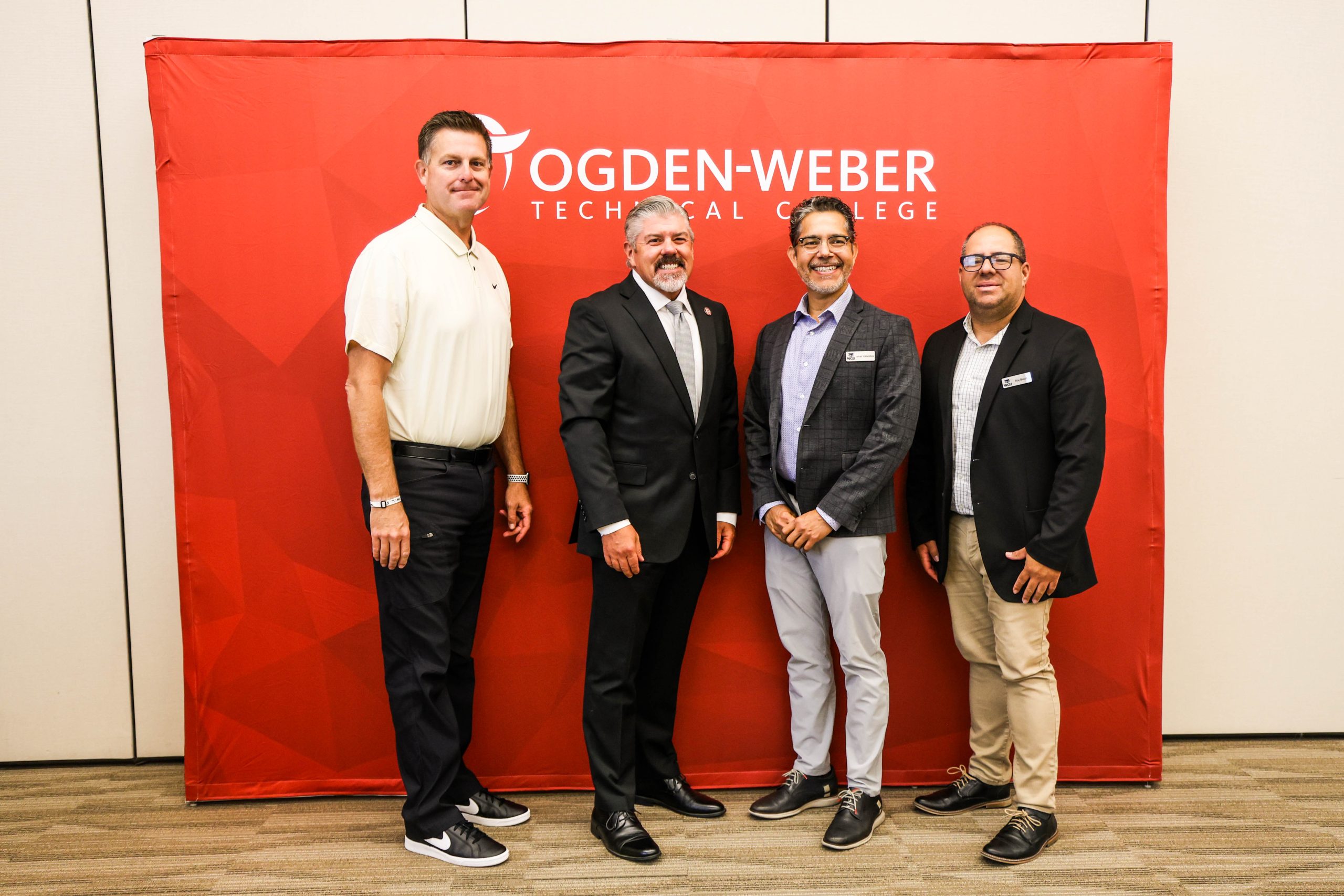 Four individuals' college leaders standing in front of a red Ogden-Weber Technical College backdrop. They are dressed in business and professional attire, posing for a photo together.
