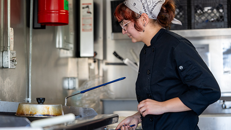 A female OTECH Culinary Arts student in a black chef coat and white bandana focuses on cooking at a commercial stovetop in a professional kitchen