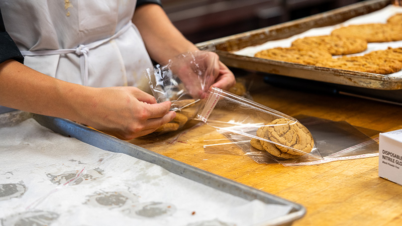An OTECH Culinary Arts student in a white apron packages freshly baked cookies into clear bags on a wooden prep table, with trays of baked goods in the background