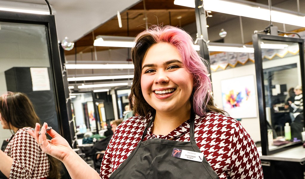 Smiling person with pink and brown hair wearing an apron in a busy salon or classroom setting