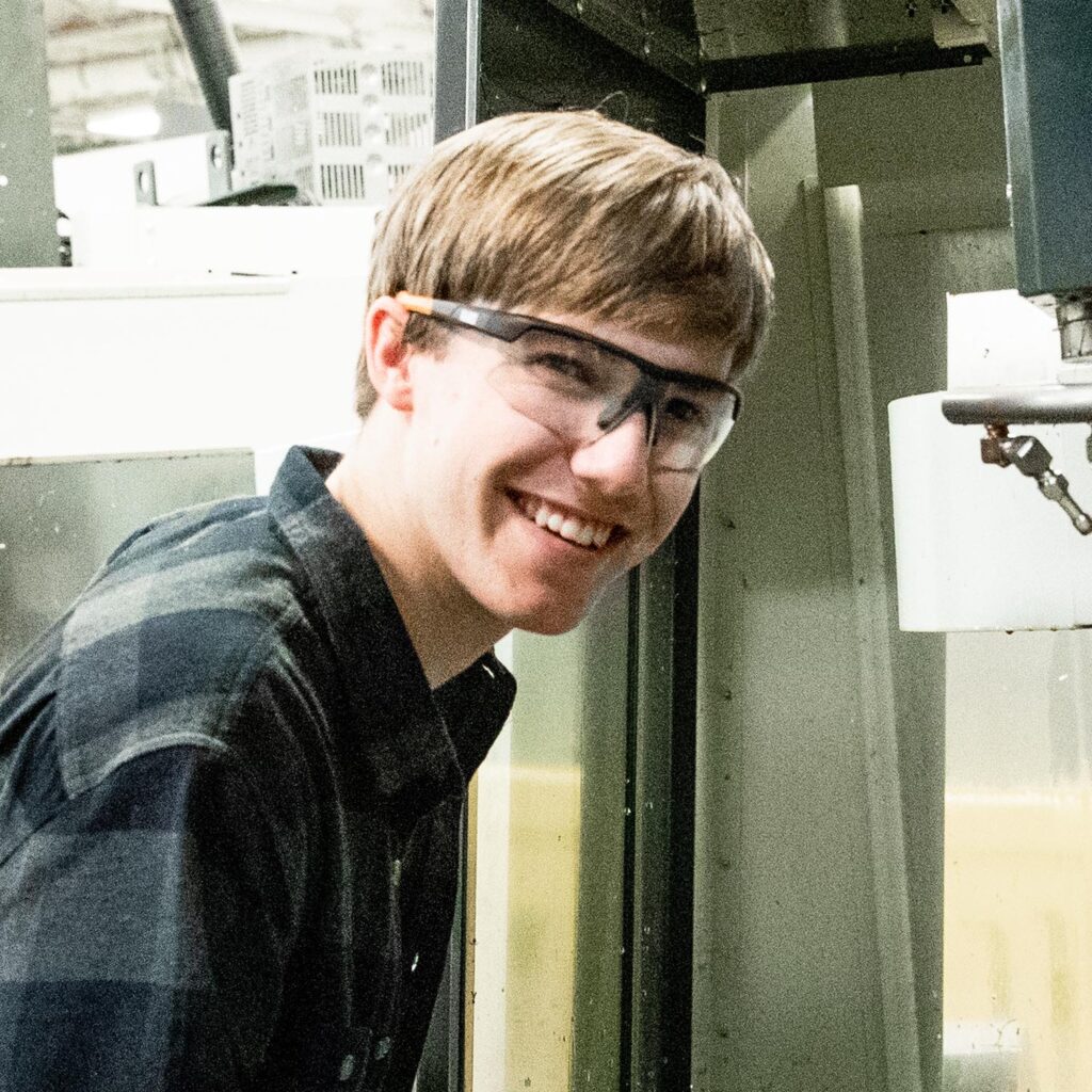 Young man in black shirt and glasses smiling in front of industrial machine.