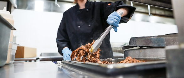 A culinary student in a black uniform and blue gloves uses tongs to prepare shredded meat on a baking tray in a commercial kitchen at OTECH