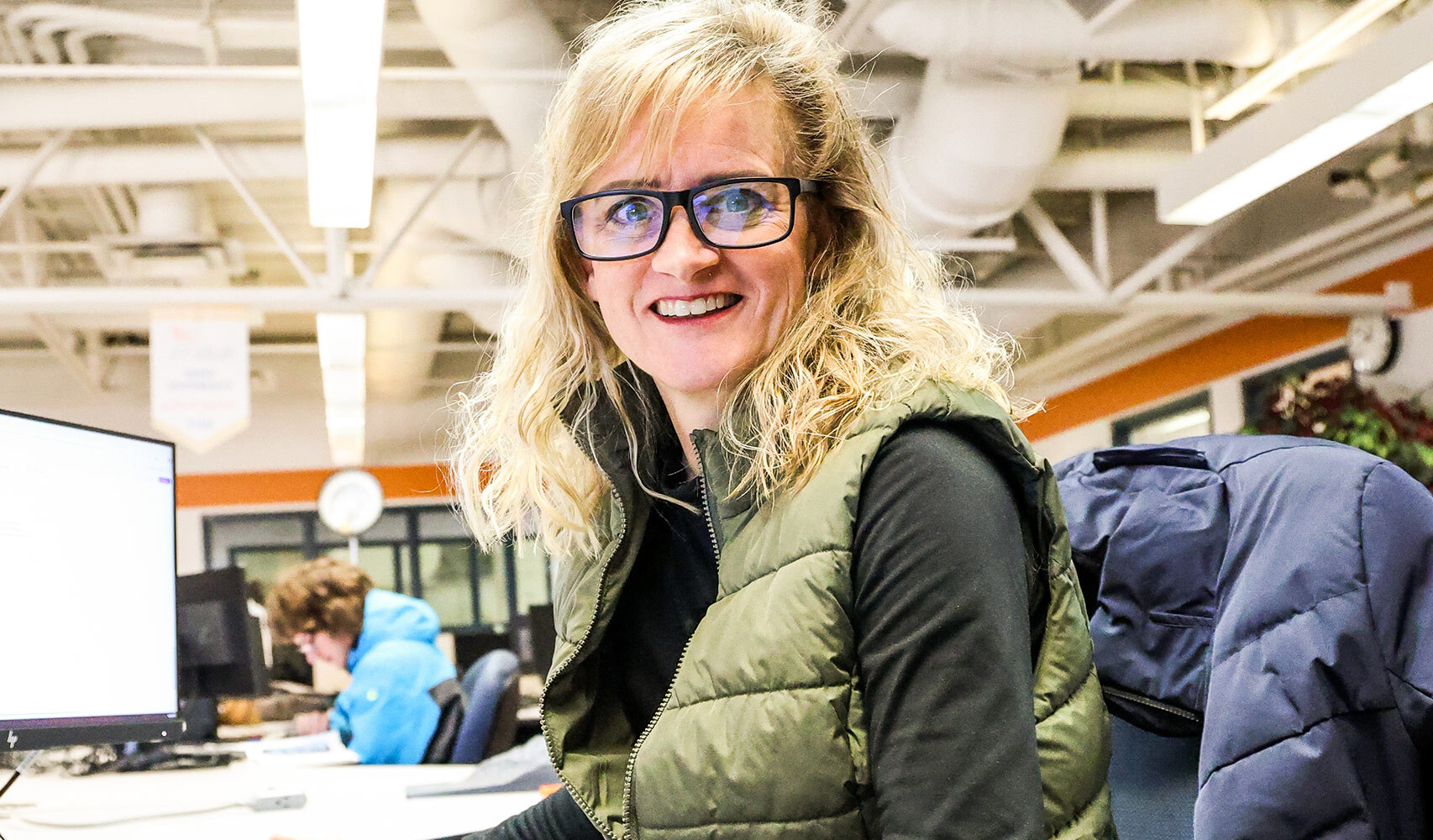 Woman with blonde hair and glasses smiling at camera in office setting.