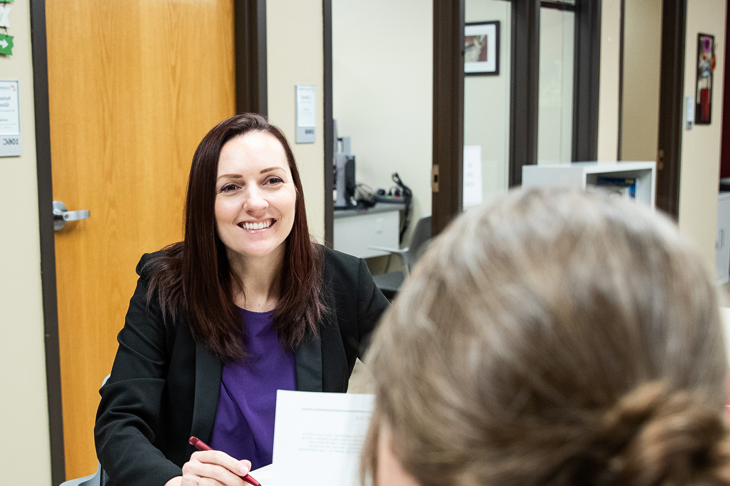 A smiling OTECH admissions advisor in a black jacket and purple top meets with a prospective student across a desk, reviewing enrollment documents during an advising appointment