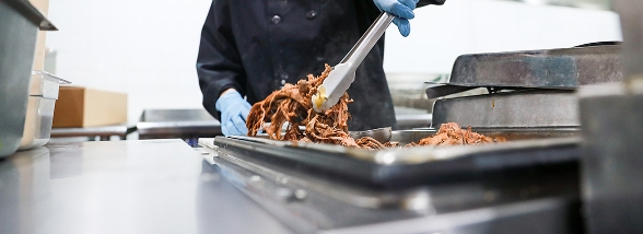 A culinary student in chef attire and blue gloves serving food with tongs in a professional kitchen, representing the campus services available at OTECH including the student-run café, beauty studio, and tech repairs