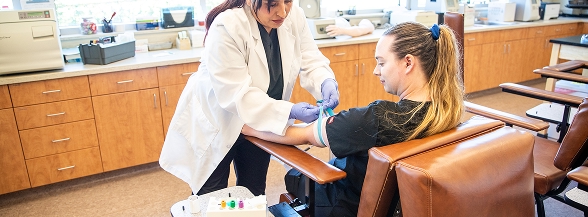 A healthcare instructor in a white lab coat performs a phlebotomy demonstration on a student's arm in a clinical training lab, showing the hands-on learning available in OTECH's community programs