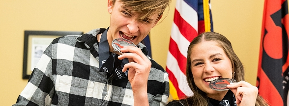 Two smiling high school students playfully bite their award medals in front of American and school flags, celebrating their achievement through OTECH's high school dual enrollment or career exploration programs
