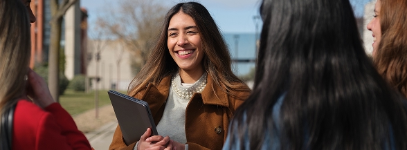 A smiling young woman holding a tablet outdoors on campus surrounded by other students, representing OTECH's community outreach programs that bring groups to campus or have OTECH attend community events