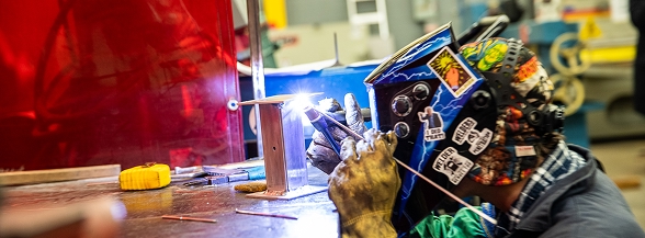 A welder wearing a decorated welding helmet and protective gloves works at a welding station with bright sparks flying behind a red safety shield, representing skilled trades workforce training available through OTECH's community programs