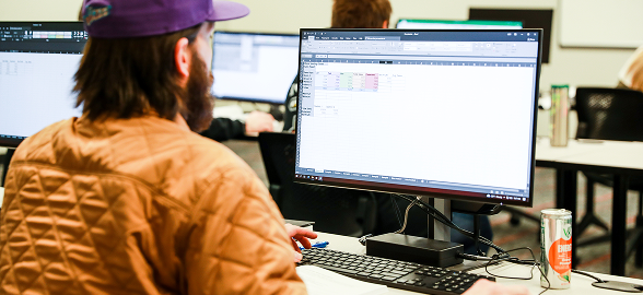 custom-fit-training-student - Ogden Weber Technical College A student viewed from behind wearing a tan jacket and purple cap works at a computer workstation displaying a spreadsheet, representing the type of custom-fit workforce training OTECH provides to employer partners