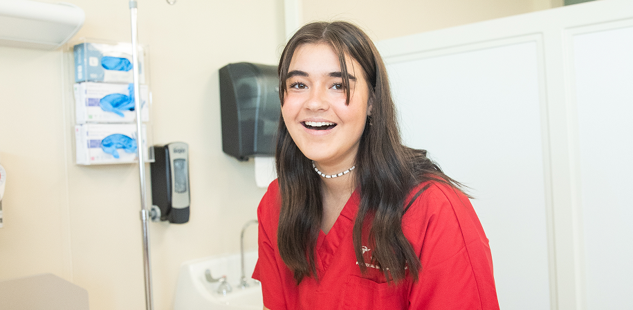A teenage girl in red scrubs laughs and smiles in a clinical training room during an OTECH summer camp healthcare experience, with medical equipment visible in the background