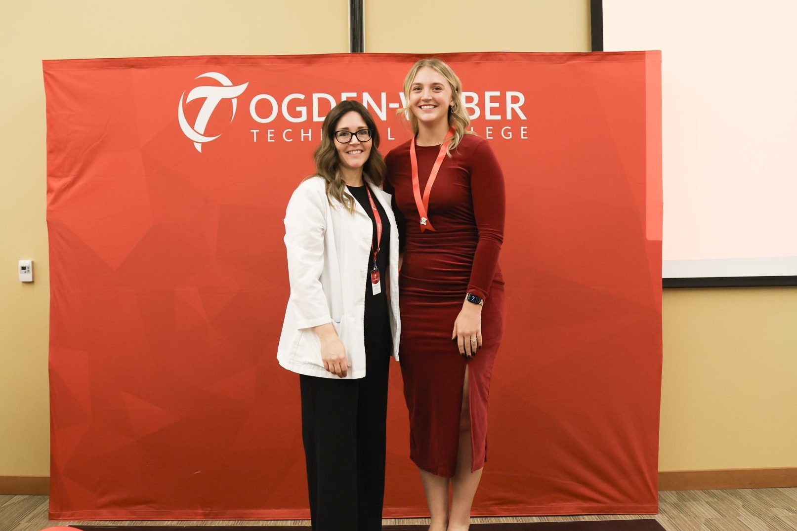 - Ogden Weber Technical College Practical nursing graduate and nursing instructor pose side in front of a red "Ogden-Weber Technical College" backdrop.