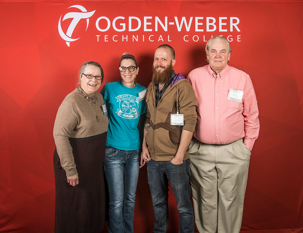 Four smiling people pose together in front of a red Ogden-Weber Technical College branded backdrop at an OTECH event