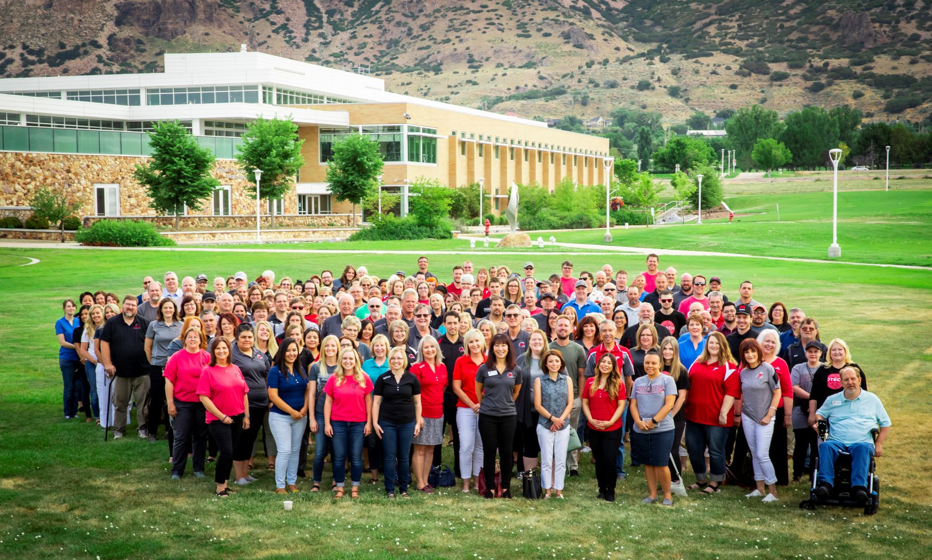 A large group of OTECH faculty and staff pose together on the front lawn of the Ogden-Weber Technical College campus with the main building and the Wasatch Mountains visible in the background