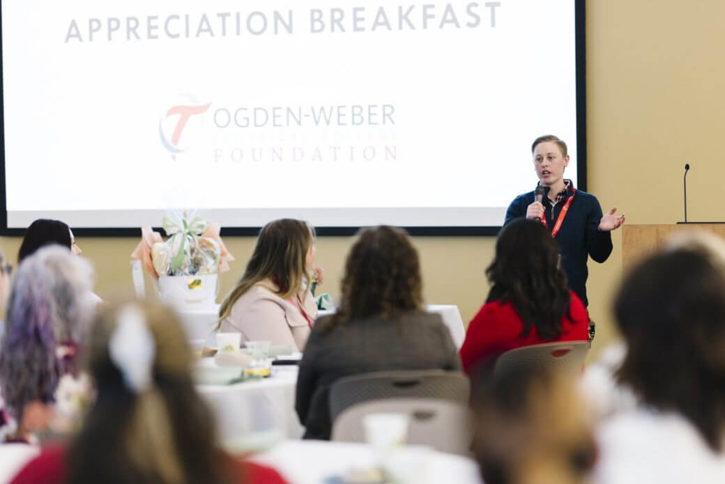 Speaker addressing attendees at OTECH’s Donor Appreciation Breakfast, with guests seated at round tables.