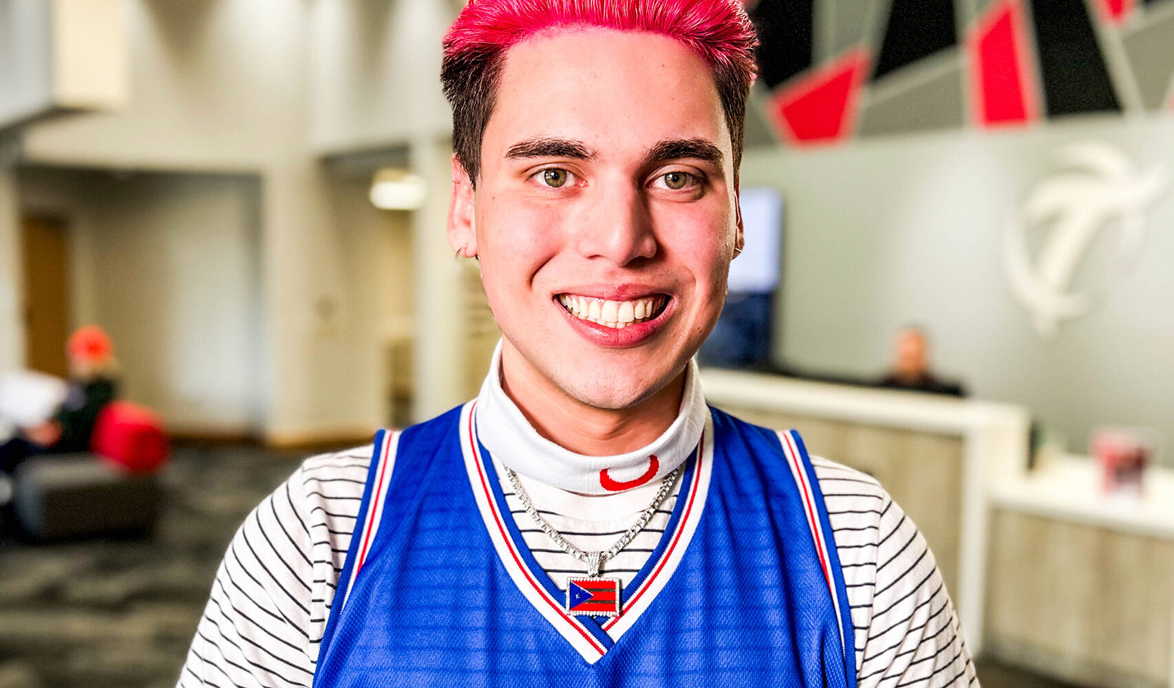 Young man with pink hair smiling at camera, wearing blue and white striped jersey with red heart design.