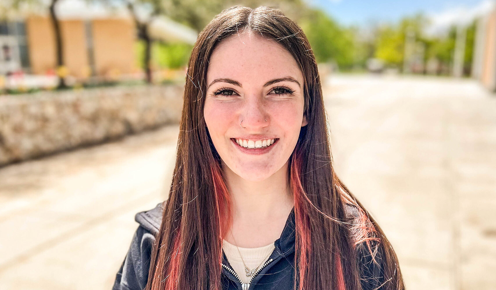 Young woman with dark hair smiling in front of a building on a sunny day.