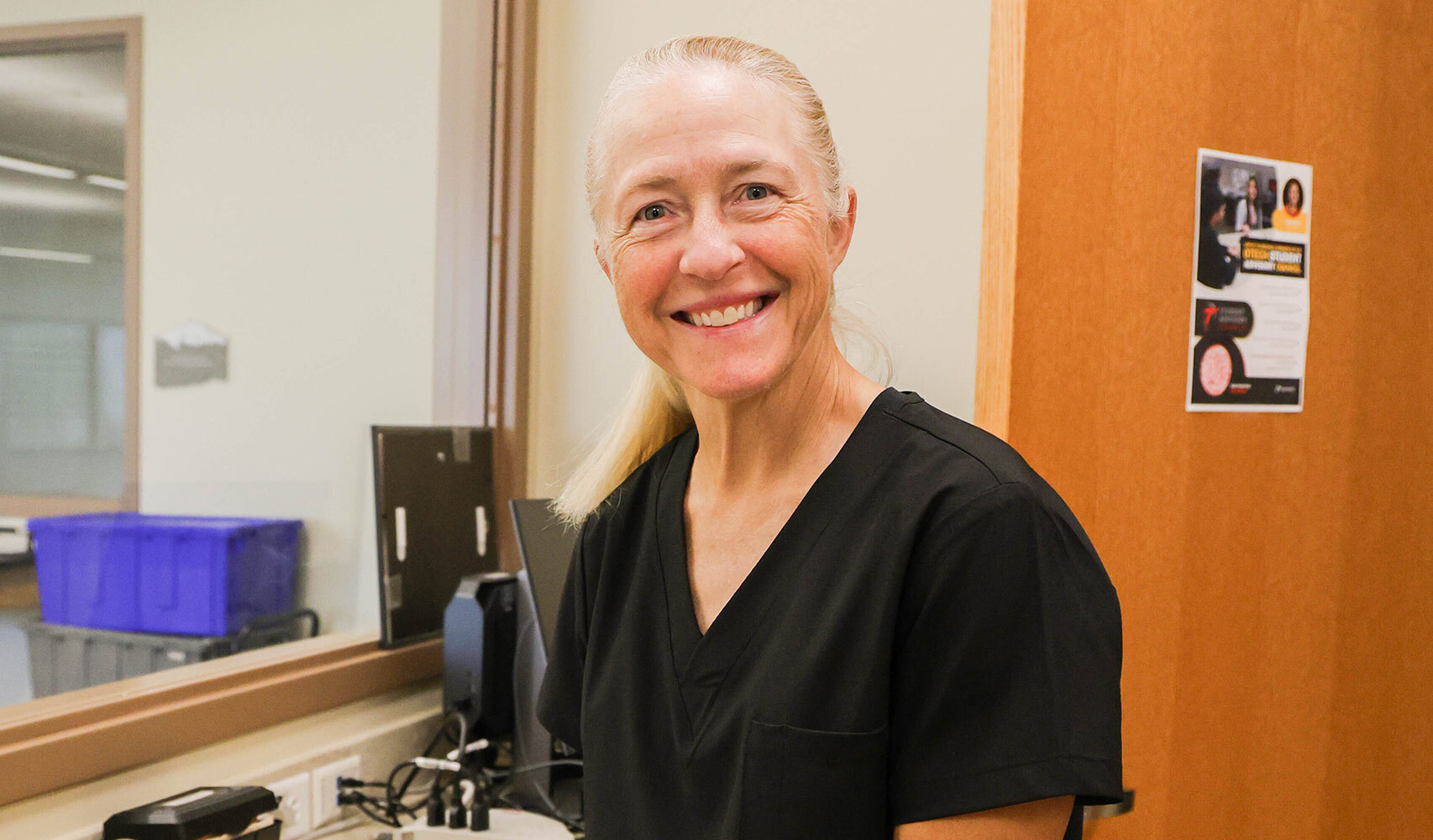 A smiling woman in black scrubs stands in a well-lit room with a computer and wooden door.