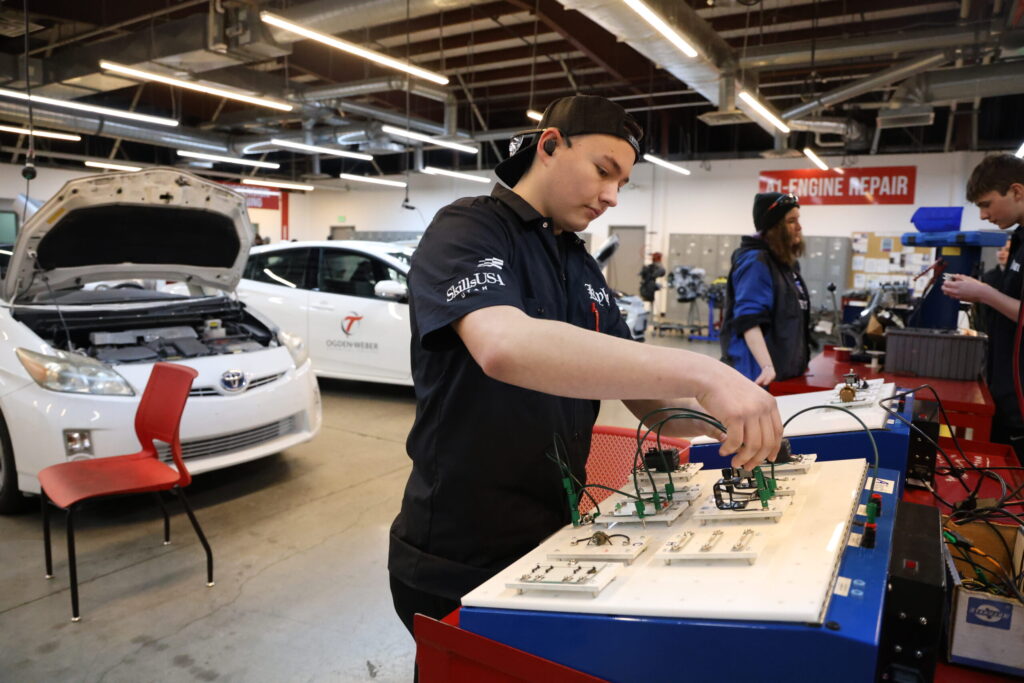 Young man in a backward-cap and black shirt wires electrical panels at a red workbench in an auto shop, with a car under repair in the background.