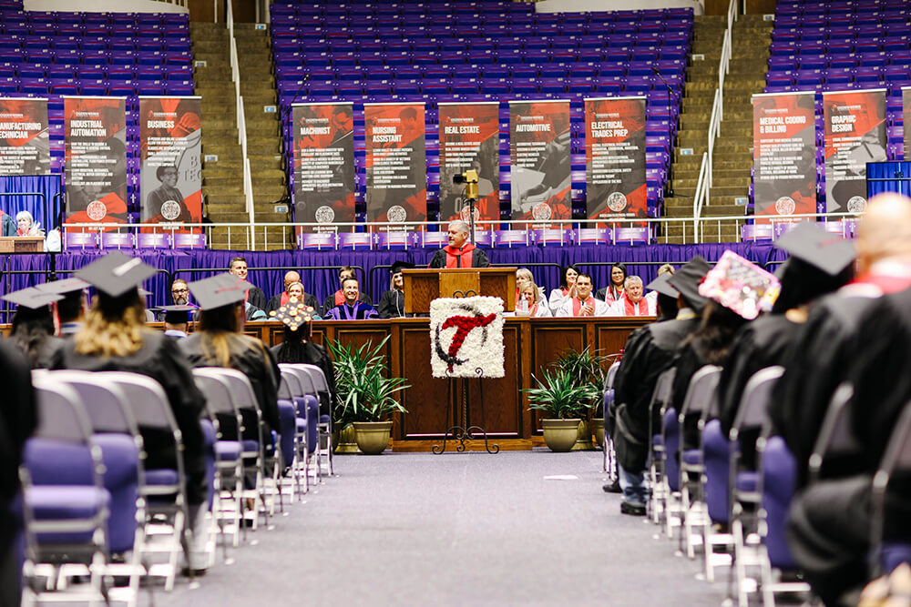 Graduates in caps and gowns sit facing a stage during a college commencement ceremony.