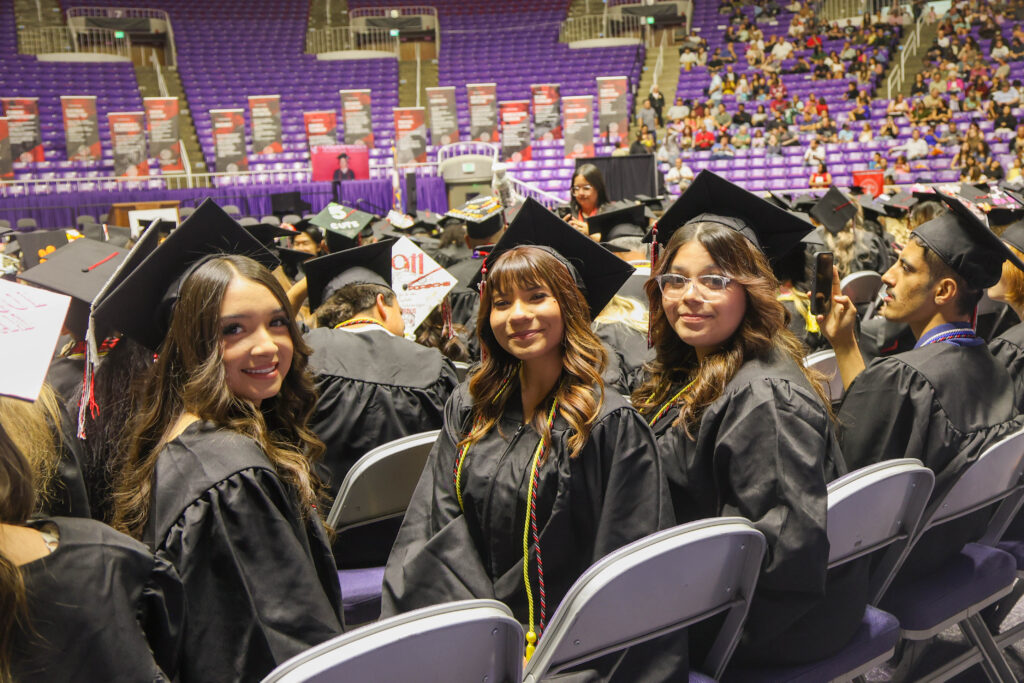 Graduates in black gowns and mortarboards seated in a stadium during a graduation ceremony, with purple seats in the background.