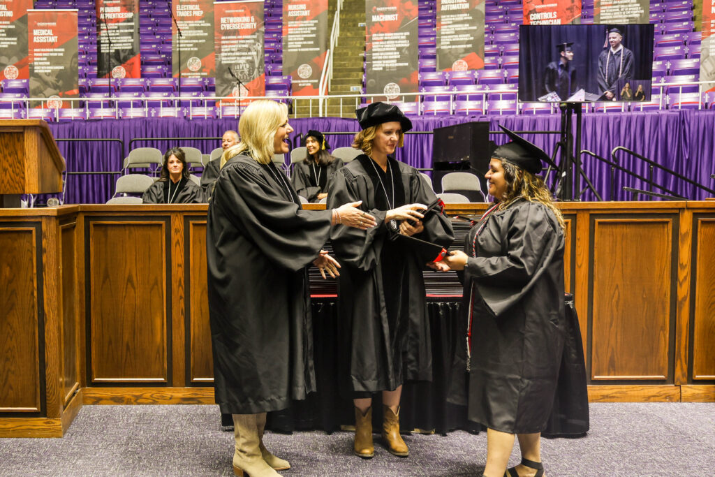 Graduates in black caps and gowns exchange a handshake/diploma in a gymnasium-style auditorium with purple seats and banners in the background.