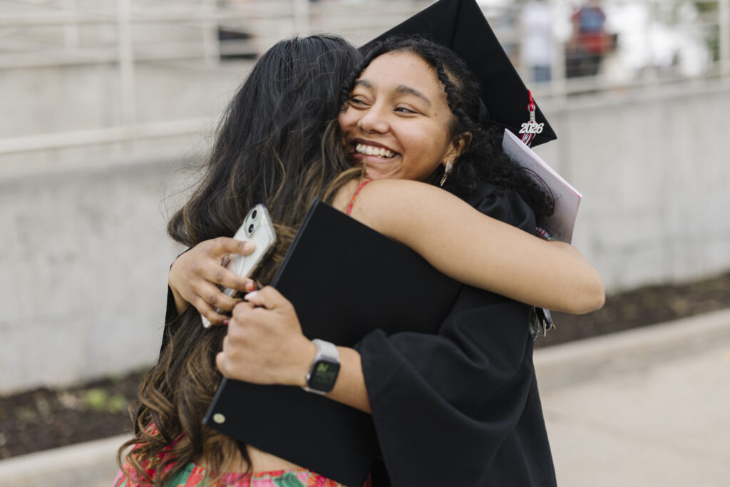 Graduate in cap and gown embraces a friend outdoors, smiling after a graduation ceremony.