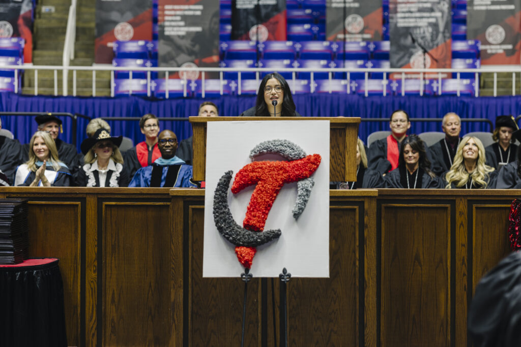 Speaker at a wooden podium addresses a graduation ceremony, with faculty in gowns seated behind and a decorative banner featuring a red ribbon in the foreground.