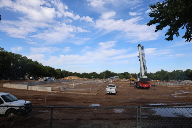 A large crane and construction vehicles at the Wasatch Peaks Pathways building site, with foundation walls and underground infrastructure visible across the work area.
