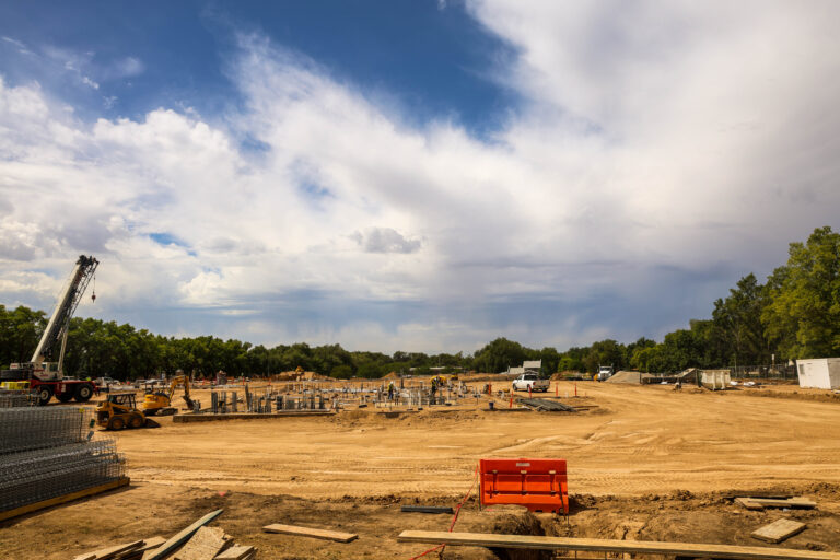Wide view of construction underway for OTECH’s Wasatch Peaks Pathways building, with a crane, construction equipment, workers, and foundation structures beneath a cloudy sky.