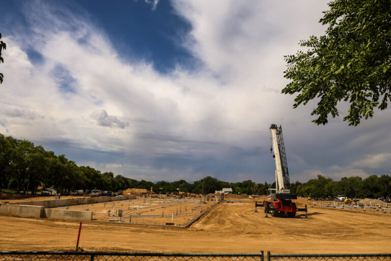 A large crane stands at the Wasatch Peaks Pathways construction site, where foundation walls, utility lines, and construction equipment are visible beneath a dramatic cloudy sky.