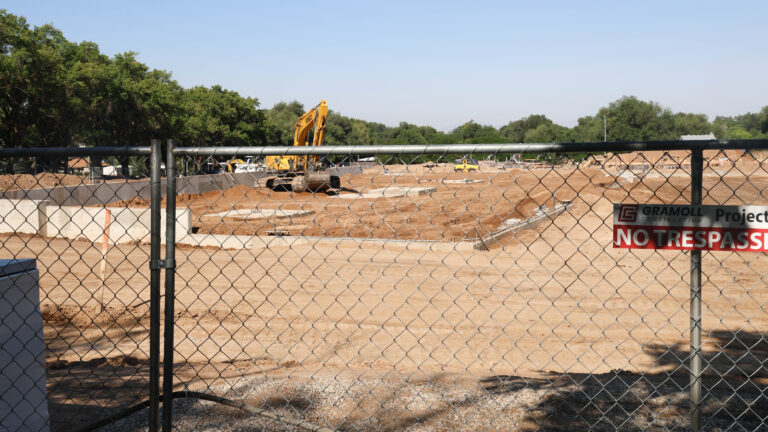 An excavator works at the Wasatch Peaks Pathways construction site behind a chain-link fence, surrounded by dirt, trenches, and concrete foundation walls.