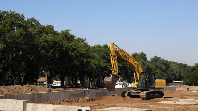 Yellow excavator at a construction site surrounded by dirt, concrete foundation walls, and mature trees.