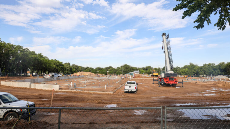 “Construction site viewed through a chain-link fence, with an excavator, concrete foundations, and trenches,