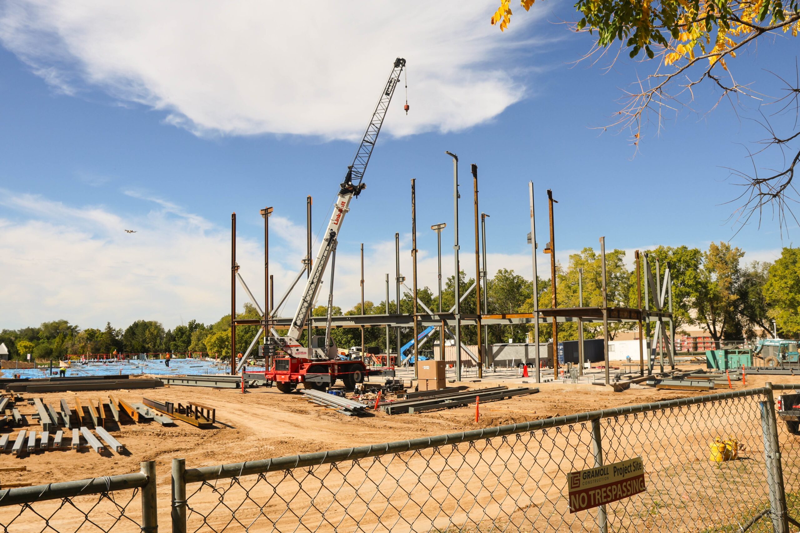 October construction progress on the new building, showing a large poured concrete foundation, steel beams staged across the site, and the first structural steel columns being installed.