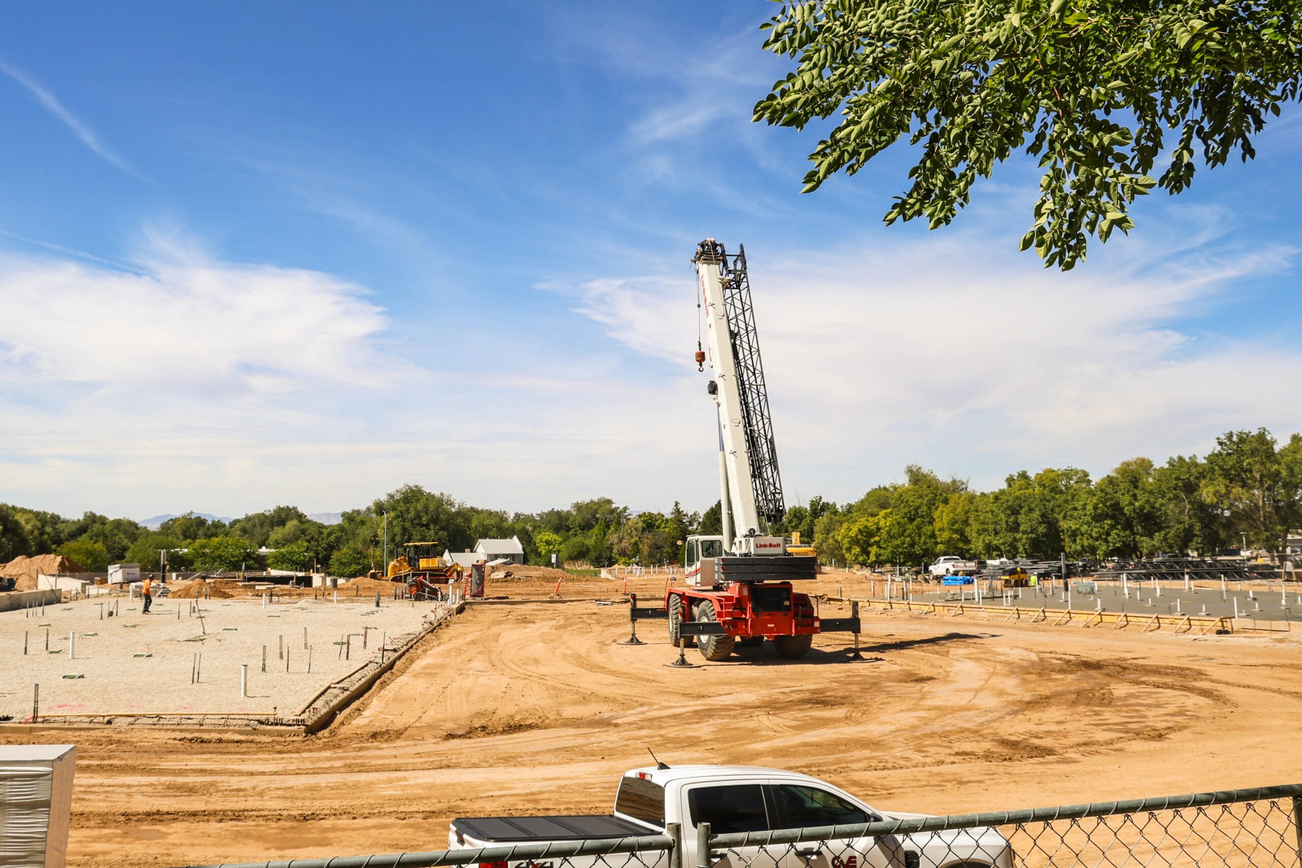 September construction progress on the new building, showing foundation work underway, utility piping throughout the site, and a large crane positioned near the future structure.